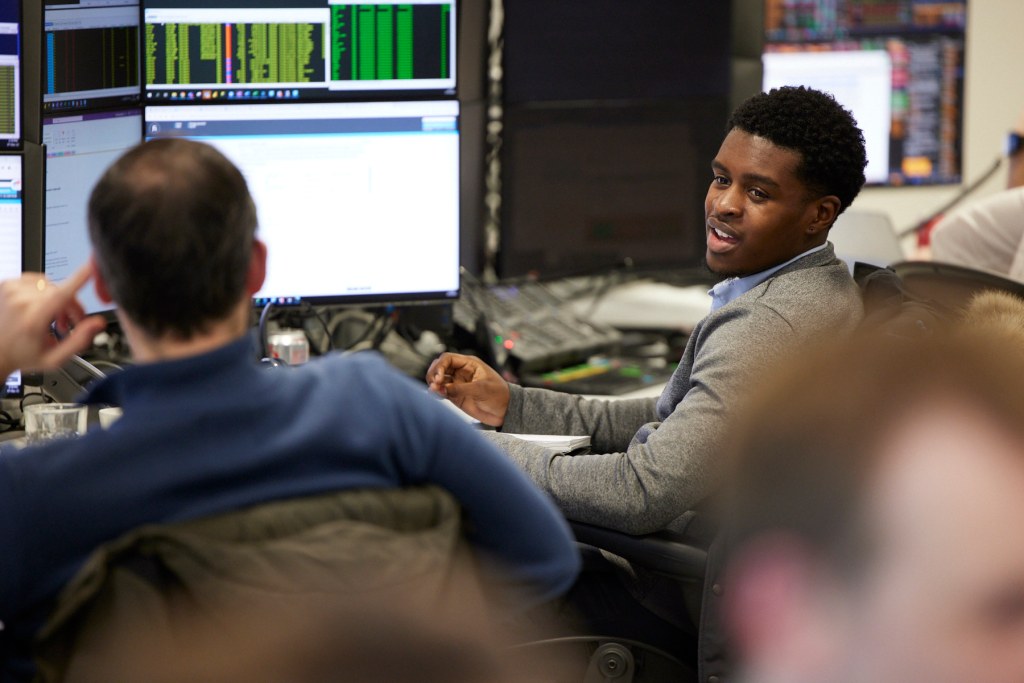 Black male banker working with other bankers in front of computer monitor in an open office.